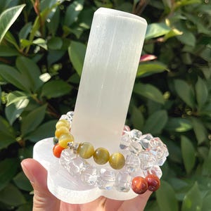 May include: A selenite crystal display stand holds two beaded bracelets. One bracelet features clear, faceted beads, while the other has yellow, orange, and brown gemstone beads. The stand is on a white, flower-shaped base.