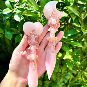 May include: Two pink crystal wands with intricate wire detailing. One wand features a heart-shaped top, the other a crescent moon. Both have a pointed base and are set against a backdrop of green foliage.
