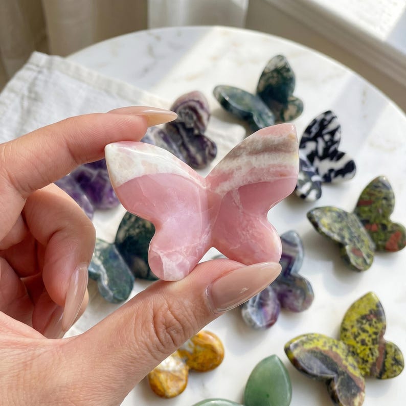 May include: A pink and white gemstone butterfly is held in a hand, with other butterfly figurines in various colors and patterns in the background. The butterflies are made of different types of stone, including green, purple, and black and white.
