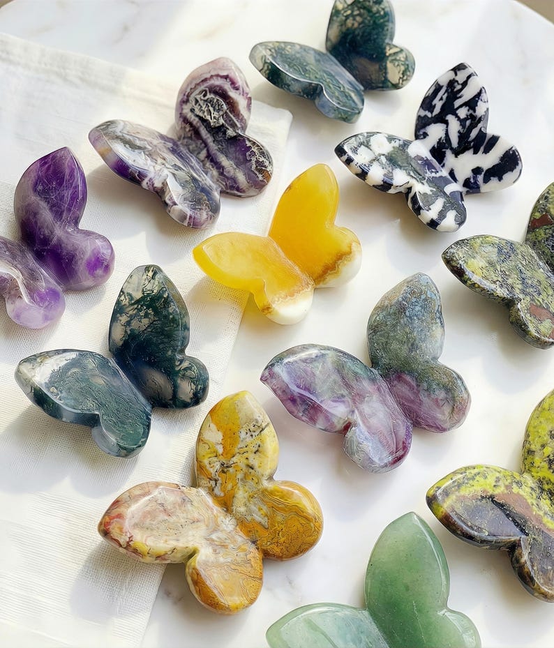 May include: A collection of gemstone butterfly figurines in various colors and patterns. The butterflies are carved from materials like amethyst, yellow quartz, and green stone. The wings display unique veining and textures, creating a natural and artistic aesthetic.