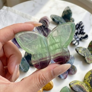 May include: A hand holds a translucent butterfly-shaped stone carving. The stone is primarily green with purple and white veining. Other butterfly carvings in various colors and patterns are visible in the background.