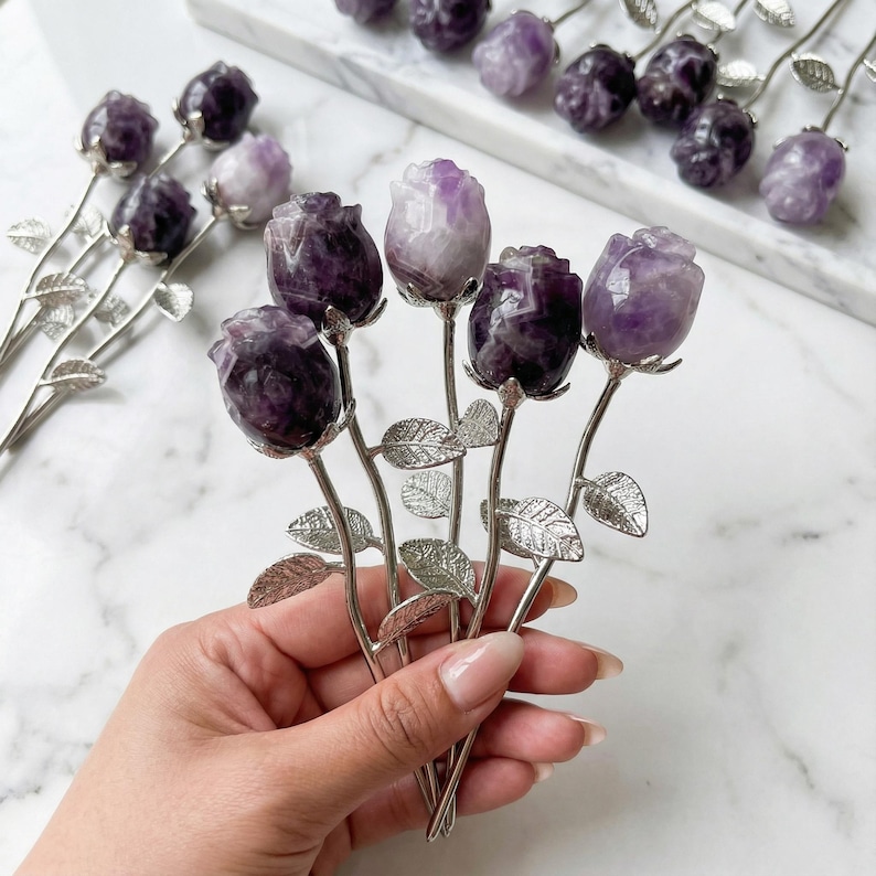 Peut inclure: Un bouquet de sculptures de roses en am&eacute;thyste. Chaque rose est sculpt&eacute;e dans une pierre pr&eacute;cieuse violette et blanche, avec des p&eacute;tales d&eacute;taill&eacute;s. Les tiges et les feuilles sont en m&eacute;tal argent&eacute;. Plusieurs bouquets similaires sont dispos&eacute;s sur une surface en marbre blanc.