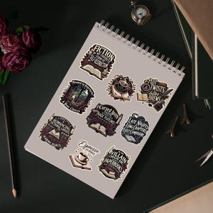 Op de afbeelding: Een verzameling stickers met gotische en literaire thema's. De stickers bevatten zinnen als "Fiction", "Cursed Taste" en "Late Nights". De stickers zijn gerangschikt op een spiraalgebonden notitieboek, met een potlood, bloemen en andere decoratieve items.