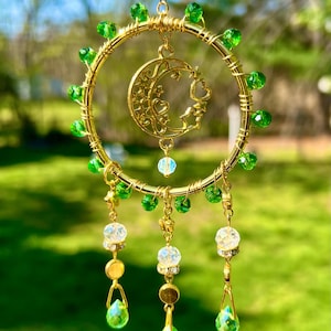 May include: A gold wire dreamcatcher with green beads and a crescent moon charm with hearts and stars. Three crystal drops hang from the bottom.