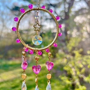 May include: A gold wire dreamcatcher with pink beads and crystal accents. The dreamcatcher has a star charm and three strands of pink heart beads with crystal drops.