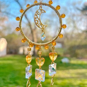 May include: A gold wire dreamcatcher with a crescent moon charm and amber beads. The dreamcatcher has three strands of crystals and gold hearts hanging from it.
