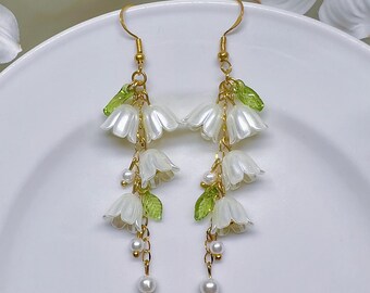 May include: A pair of gold dangle earrings with white flower charms and green leaf accents. The earrings feature a chain with small white pearl beads.