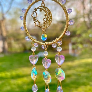 May include: A gold wire dreamcatcher with a crescent moon and stars design. The dreamcatcher has lavender beads and iridescent crystal accents. Three strands of iridescent crystals hang from the bottom of the dreamcatcher.