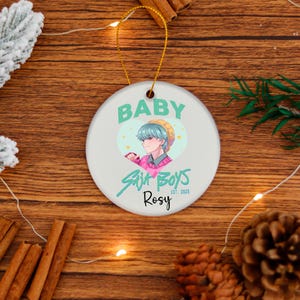 Peut inclure: Une décoration de Noël ronde et blanche avec une ficelle dorée. L'ornement porte le texte "BABY" au-dessus d'une illustration d'une personne aux cheveux bleus et le texte "Suit Boys Rosy Est. 2025".