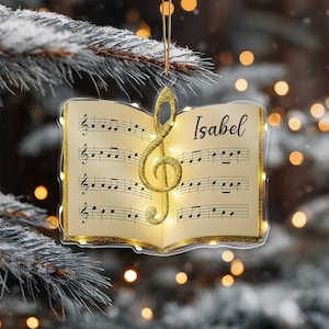 May include: A Christmas ornament shaped like an open music book with musical notes and a golden treble clef. The name "Isabel" is written on the right page. The ornament is trimmed with gold glitter and has a string for hanging.