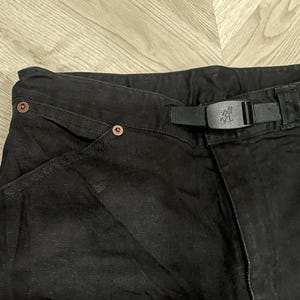 Puede incluir: Primer plano de pantalones negros con cintur&oacute;n y hebilla negros integrados. Los pantalones tienen remaches de color cobre y un dise&ntilde;o de bolsillo &uacute;nico. La hebilla del cintur&oacute;n tiene un logotipo. La tela parece ser un material tejido duradero.