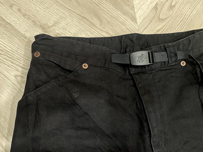 Puede incluir: Primer plano de pantalones negros con cintur&oacute;n y hebilla negros integrados. Los pantalones tienen remaches de color cobre y un dise&ntilde;o de bolsillo &uacute;nico. La hebilla del cintur&oacute;n tiene un logotipo. La tela parece ser un material tejido duradero.