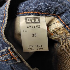 May include: Close-up of blue denim jeans with a tag that reads "EDWIN" and "421XVS". The tag also indicates a size of "36". The jeans have orange stitching and a classic denim look.