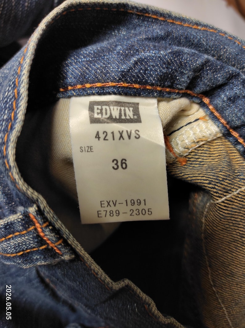 May include: Close-up of blue denim jeans with a tag that reads "EDWIN" and "421XVS". The tag also indicates a size of "36". The jeans have orange stitching and a classic denim look.