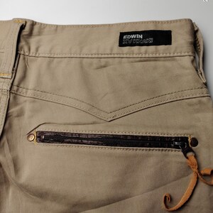 May include: Close-up of khaki-colored pants with a zippered pocket and a leather accent. The pants feature the "EDWIN XV KHAKI" label. The zipper pull has a leather strap attached. The stitching details are visible.
