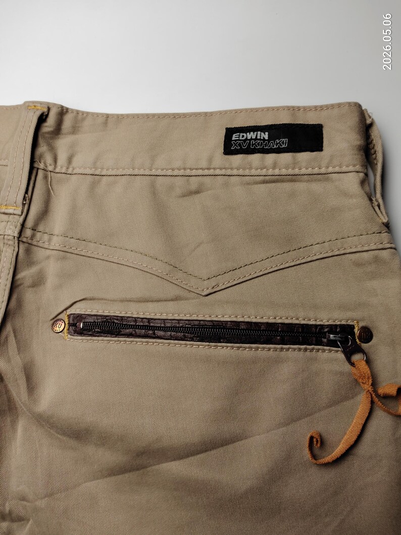May include: Close-up of khaki-colored pants with a zippered pocket and a leather accent. The pants feature the "EDWIN XV KHAKI" label. The zipper pull has a leather strap attached. The stitching details are visible.