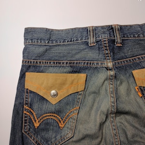 May include: Close-up of the back of blue denim jeans. The jeans feature a light brown suede pocket with orange stitching and a silver button. The back pocket has an orange embroidered design. The jeans are faded.