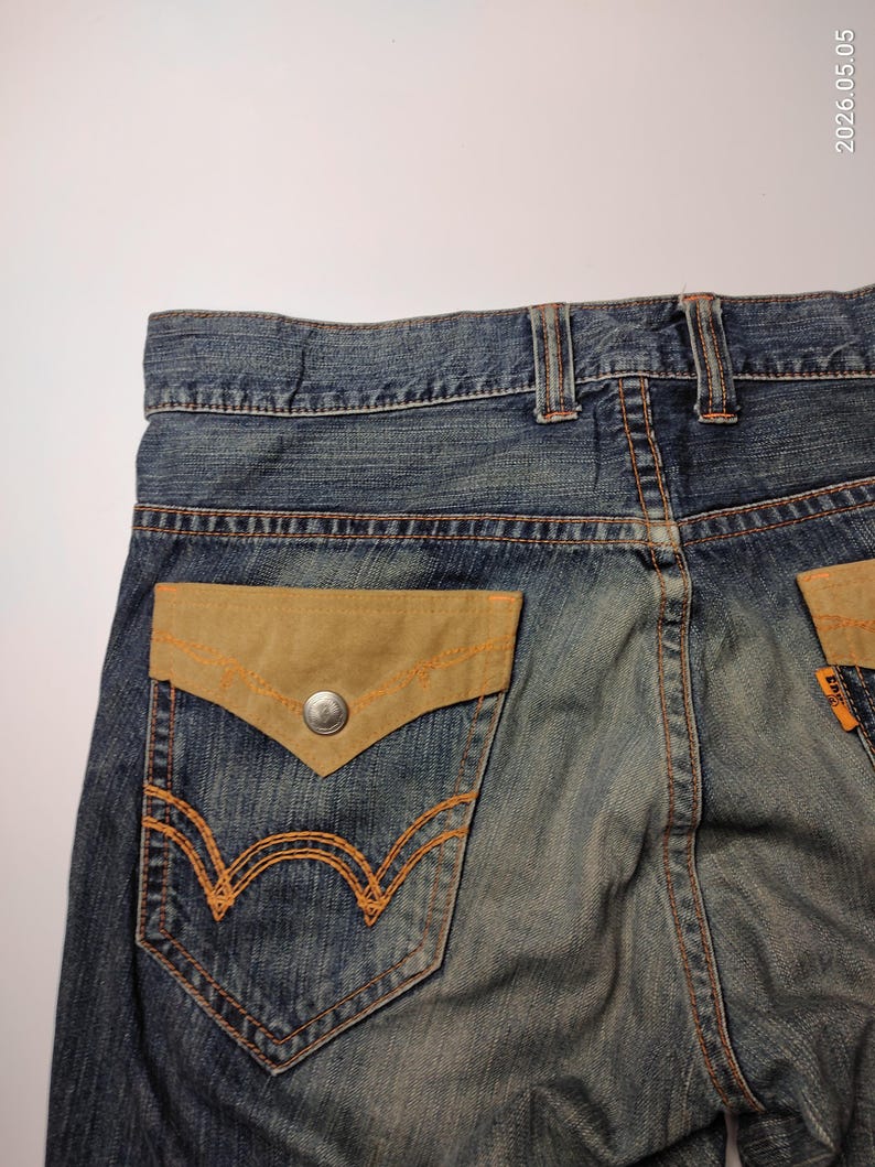May include: Close-up of the back of blue denim jeans. The jeans feature a light brown suede pocket with orange stitching and a silver button. The back pocket has an orange embroidered design. The jeans are faded.