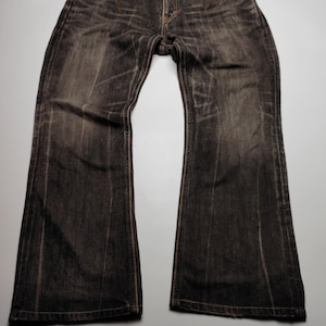 May include: Dark wash denim jeans with a flared leg. The jeans have a faded appearance with brown stitching and pocket accents. The waistband is a lighter color, and the jeans are laid flat on a light gray surface.