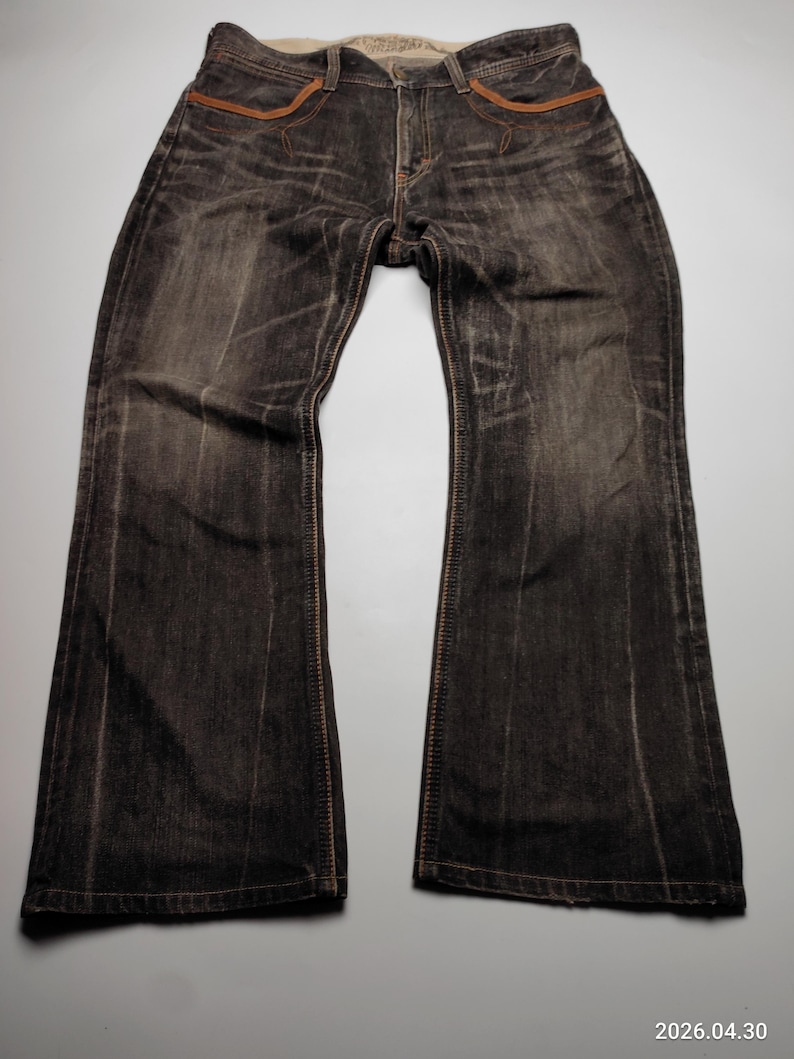 May include: Dark wash denim jeans with a flared leg. The jeans have a faded appearance with brown stitching and pocket accents. The waistband is a lighter color, and the jeans are laid flat on a light gray surface.