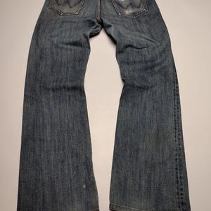 Peut inclure: Jean en denim bleu d&eacute;lav&eacute; avec une conception classique &agrave; cinq poches. Le jean pr&eacute;sente le logo Wrangler sur les poches arri&egrave;re et un aspect us&eacute;, sugg&eacute;rant un style vintage. Le jean est pos&eacute; &agrave; plat, mettant en valeur toute la longueur et les d&eacute;tails.