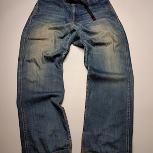 May include: Blue denim trousers with a black belt. The trousers have a relaxed fit and a faded wash, with visible wear marks. The belt has a red accent. The trousers are laid flat on a white surface.