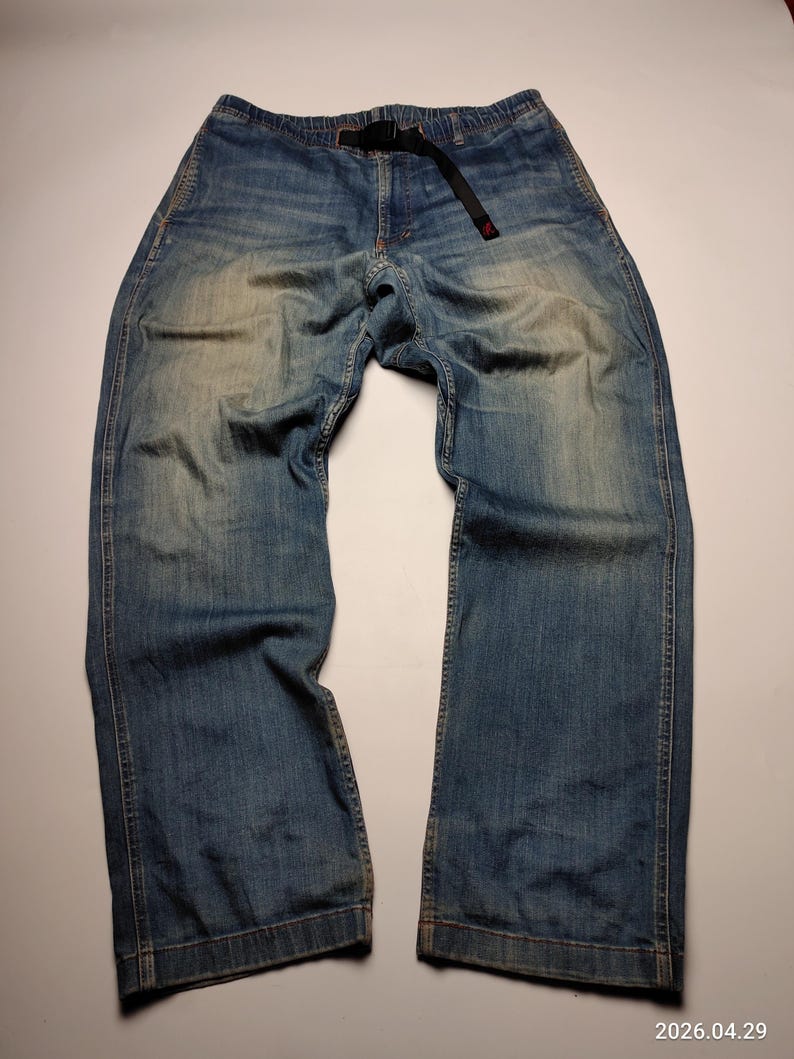 May include: Blue denim trousers with a black belt. The trousers have a relaxed fit and a faded wash, with visible wear marks. The belt has a red accent. The trousers are laid flat on a white surface.