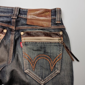 Puede incluir: Primer plano de vaqueros de mezclilla azules con costuras naranjas. Incluye un parche de cuero marr&oacute;n, un bolsillo con cremallera y una etiqueta roja. El bolsillo trasero tiene un dise&ntilde;o bordado naranja distintivo.