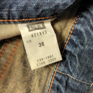 May include: Close-up of a denim garment with a white tag. The tag reads "EDWIN" above "421XVS" and "SIZE 36". Below the size, the tag displays "EXV-1991" and "E789-2305". The denim is blue with orange stitching.