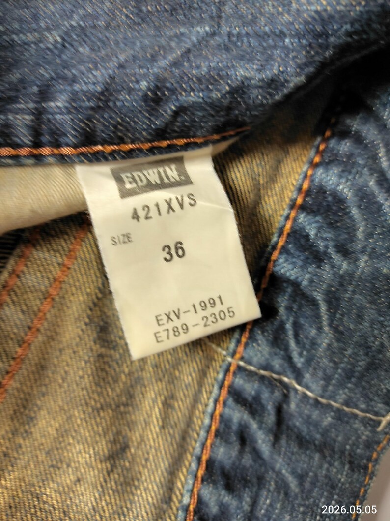 May include: Close-up of a denim garment with a white tag. The tag reads "EDWIN" above "421XVS" and "SIZE 36". Below the size, the tag displays "EXV-1991" and "E789-2305". The denim is blue with orange stitching.