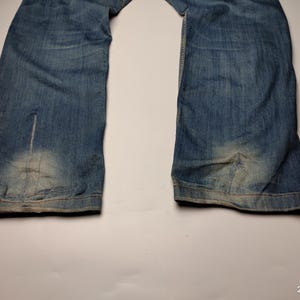May include: Faded blue denim jeans with a light wash on the lower legs. The jeans have a classic five-pocket design and a straight leg cut. The denim shows signs of wear and tear, adding to the vintage aesthetic.