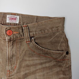 May include: Close-up of brown denim jeans with orange stitching and a red button. The jeans have a classic five-pocket design and a faded appearance. The brand name "EDWIN" is visible on a tag.