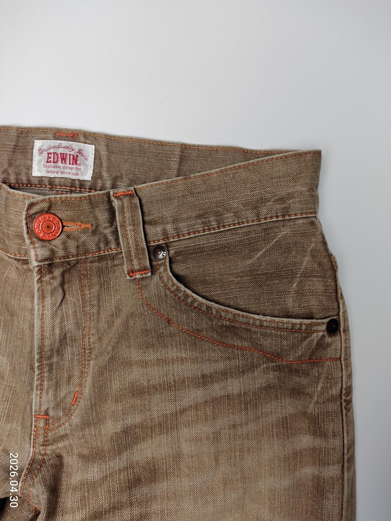 May include: Close-up of brown denim jeans with orange stitching and a red button. The jeans have a classic five-pocket design and a faded appearance. The brand name "EDWIN" is visible on a tag.