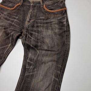 May include: Dark gray denim jeans with brown accents on the pockets and seams. The jeans have a faded, worn look and a classic five-pocket design. The waistband features a printed label.