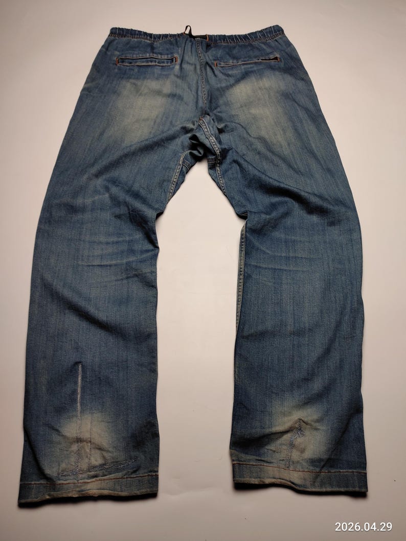 May include: Faded blue denim trousers with an elasticated waistband and drawstring. The jeans have a relaxed fit and show signs of wear, with lighter patches on the legs. The back pockets are visible, and the overall style is casual.