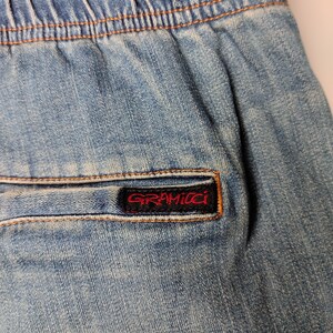 May include: Close-up of light blue denim shorts with an elasticated waistband and a back pocket. The pocket features a red and black Gramicci brand label. The shorts have visible stitching and a faded appearance.
