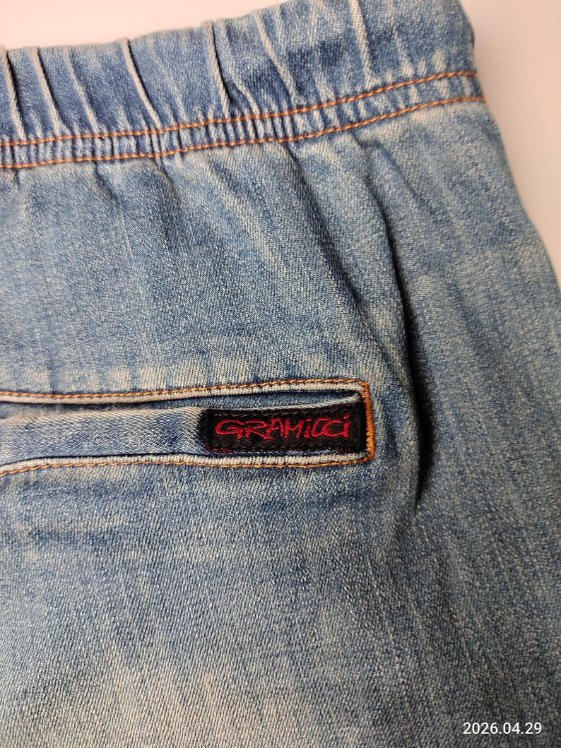 May include: Close-up of light blue denim shorts with an elasticated waistband and a back pocket. The pocket features a red and black Gramicci brand label. The shorts have visible stitching and a faded appearance.