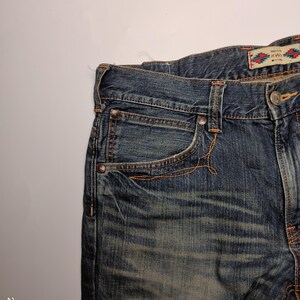 May include: Close-up of blue denim jeans with orange stitching. The jeans have a faded wash and a small decorative tag with text and a colorful pattern. The jeans have a classic five-pocket design.