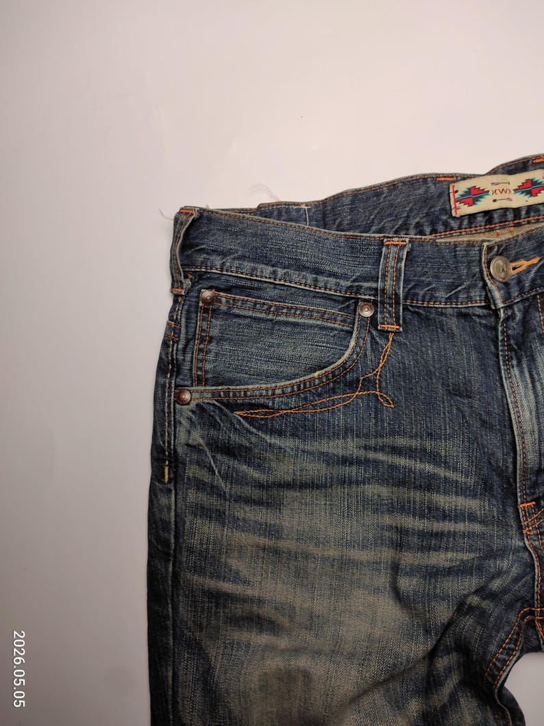 May include: Close-up of blue denim jeans with orange stitching. The jeans have a faded wash and a small decorative tag with text and a colorful pattern. The jeans have a classic five-pocket design.