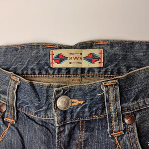 May include: Close-up of blue denim jeans with orange stitching. The jeans feature a button closure and a decorative label with a geometric design in red, turquoise, and white. The jeans are a classic style.