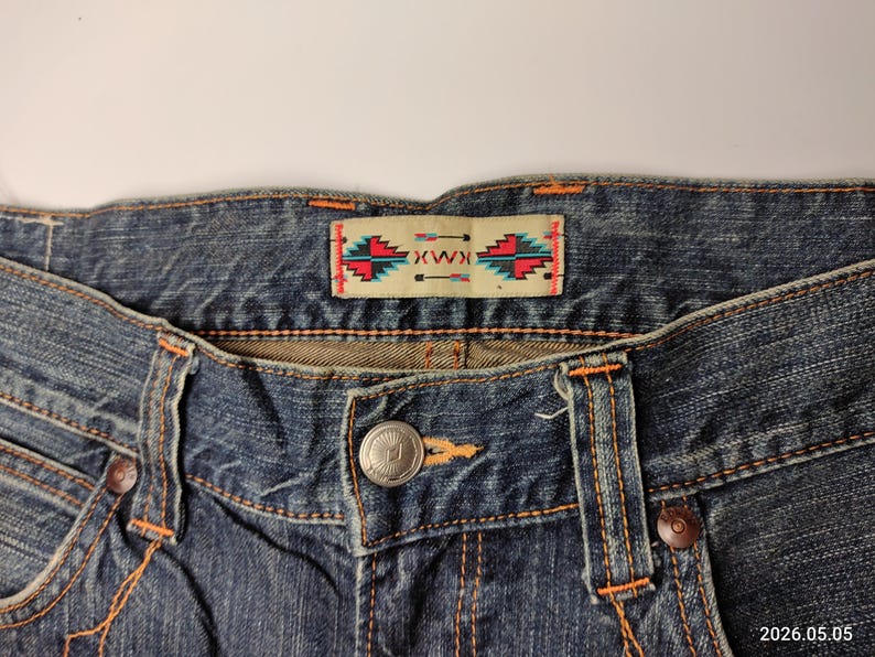 May include: Close-up of blue denim jeans with orange stitching. The jeans feature a button closure and a decorative label with a geometric design in red, turquoise, and white. The jeans are a classic style.