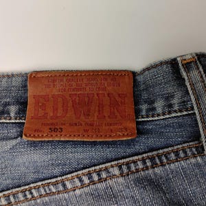 May include: Close-up of the back of blue denim jeans with a brown leather patch. The patch features the brand name "EDWIN" in red lettering, along with the text "GENUINE QUALITY JEANS FOR ALL THE PEOPLE OF THE WORLD BY EDWIN FOR CENTURIES TO COME".