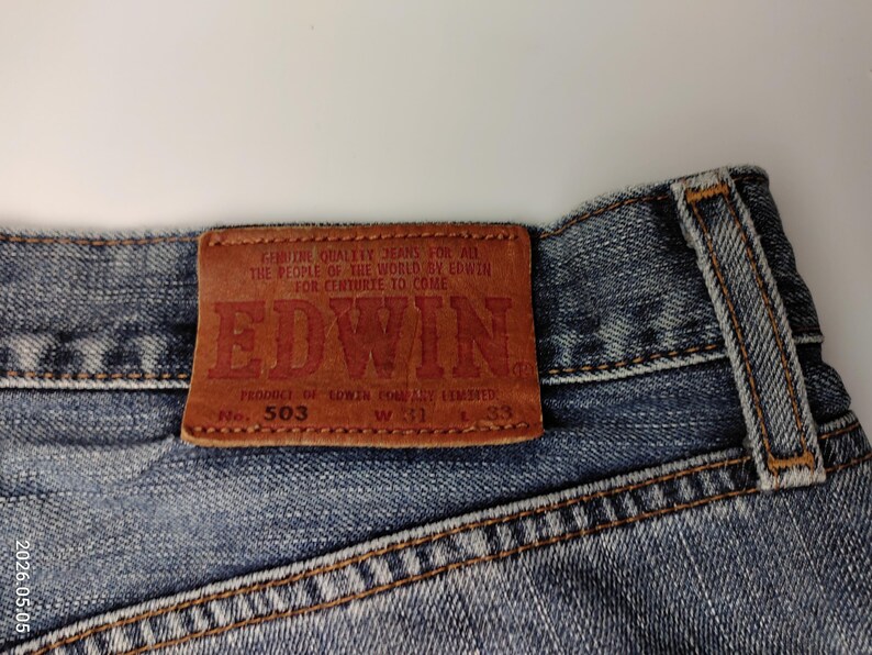 May include: Close-up of the back of blue denim jeans with a brown leather patch. The patch features the brand name "EDWIN" in red lettering, along with the text "GENUINE QUALITY JEANS FOR ALL THE PEOPLE OF THE WORLD BY EDWIN FOR CENTURIES TO COME".