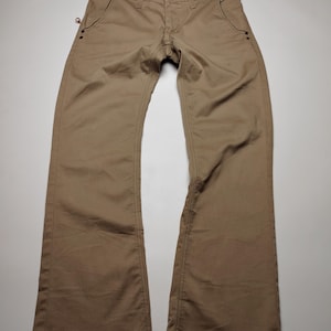 May include: Khaki-colored pants with a button and zipper closure. The pants have two front pockets and two back pockets. The stitching is visible, and the pants appear to be made of a durable material. The pants are laid flat on a white surface.