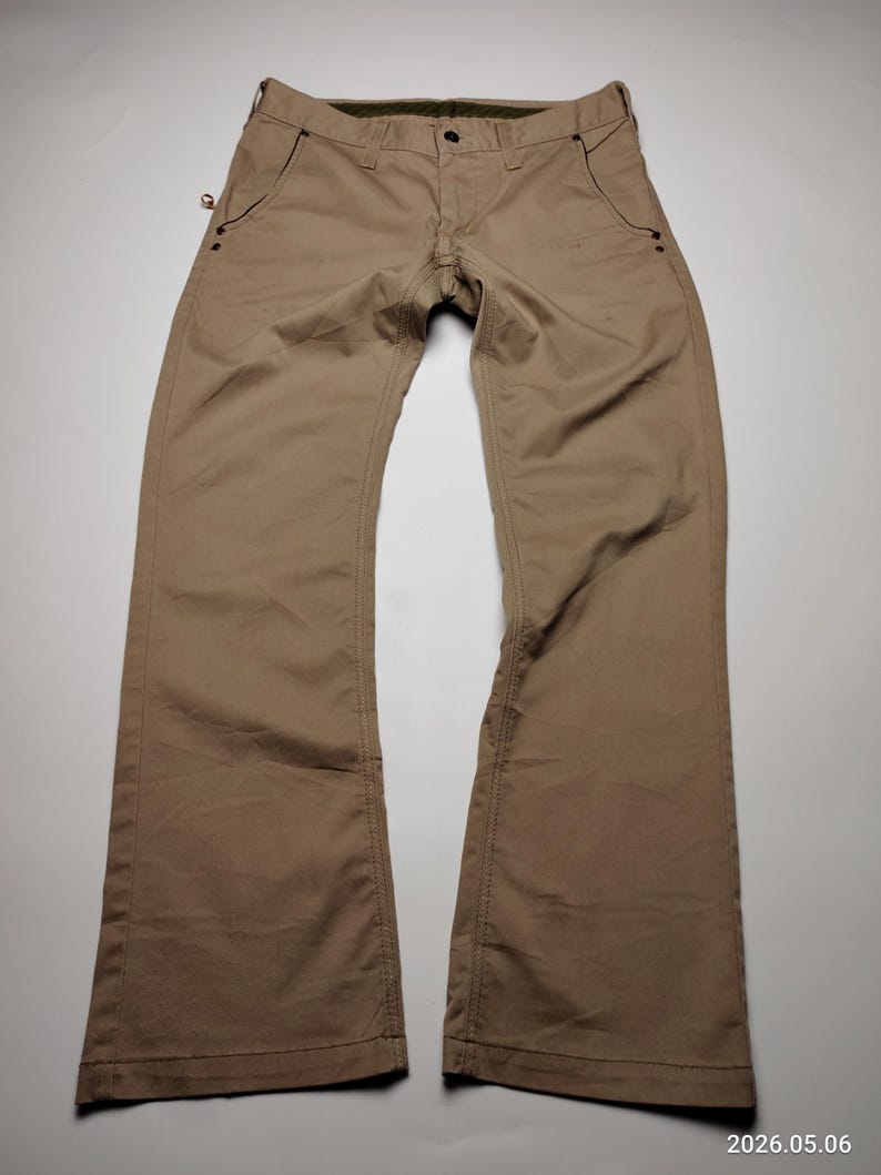 May include: Khaki-colored pants with a button and zipper closure. The pants have two front pockets and two back pockets. The stitching is visible, and the pants appear to be made of a durable material. The pants are laid flat on a white surface.