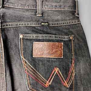 May include: Close-up of black denim jeans with the Wrangler logo on the back pocket. The pocket features a decorative stitch design in red, orange, and yellow. The jeans have a classic five-pocket design.