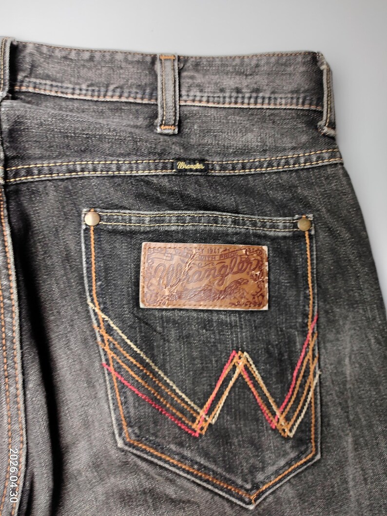 May include: Close-up of black denim jeans with the Wrangler logo on the back pocket. The pocket features a decorative stitch design in red, orange, and yellow. The jeans have a classic five-pocket design.