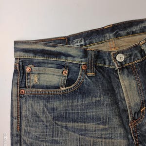 May include: Close-up of blue denim jeans with a faded wash. The jeans feature copper rivets, a button closure, and a small pocket. The brand name "EDWIN" is visible on the waistband. The denim has a worn, vintage look.