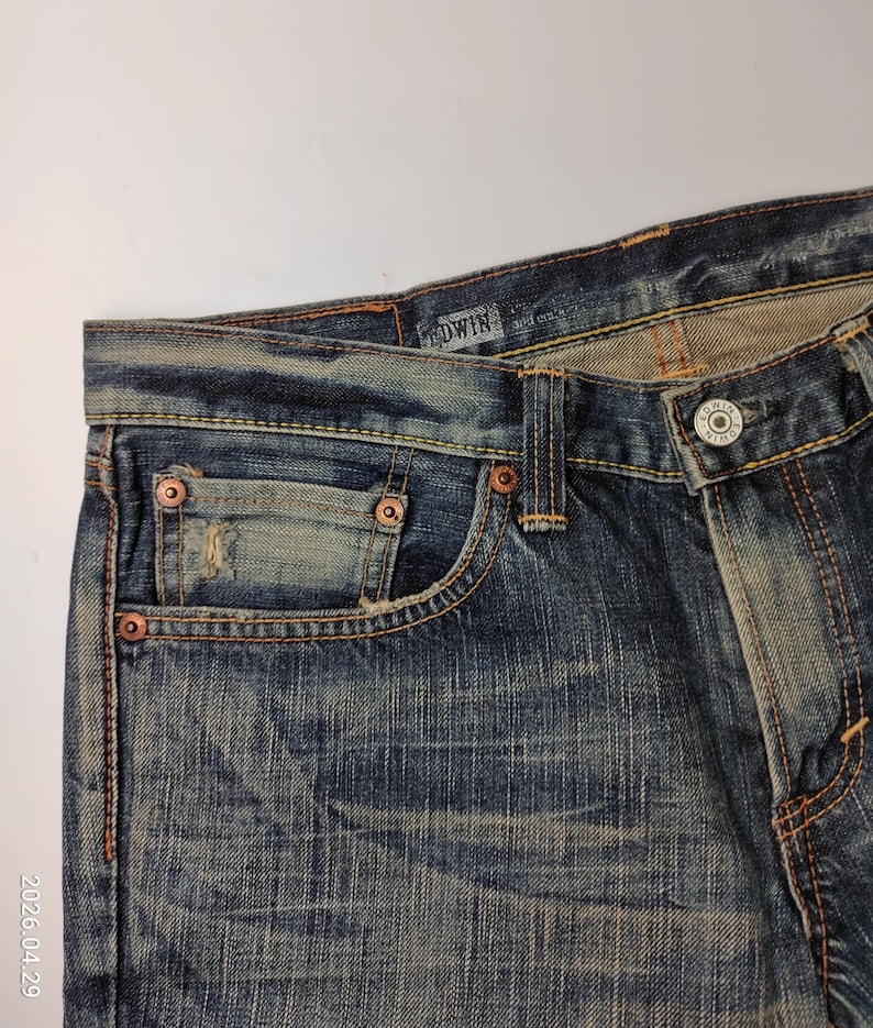 May include: Close-up of blue denim jeans with a faded wash. The jeans feature copper rivets, a button closure, and a small pocket. The brand name "EDWIN" is visible on the waistband. The denim has a worn, vintage look.