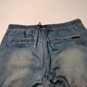 May include: Faded blue denim joggers with an elasticated waistband and drawstring. The back features two pockets with orange stitching and a small, rectangular logo patch. The denim has a worn appearance.
